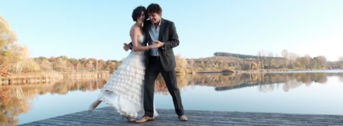 Tanzpaar im Balloutfit auf einem Steg am See. Im Hintergrund herbstliche Laubbäume. Sie steht in seinem rechten Arm das rechte Bein weggesteckt die gefassten Hände vor seinem Körper